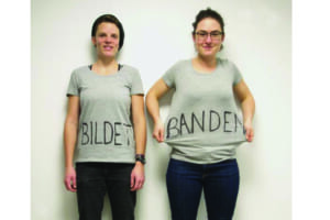 Image of two people in front of a white wall, visible up to the knees. The left person is minimally smaller than the right. The wears a black cap and short brown hair and black jeans. On her light gray T-shirt is written on the front in handwriting and small caps BILDET (create, form). Her arms hang relaxed down her body, she smiles. The person on the right has longer hair tied back and black round glasses and blue jeans. She is wearing the same T-shirt and is pulling her sides apart at the waist with her two arms. The T-shirt says in this area, also handwritten in large black letters: BANDEN (gangs, groups). She also looks into the camera and smiles.