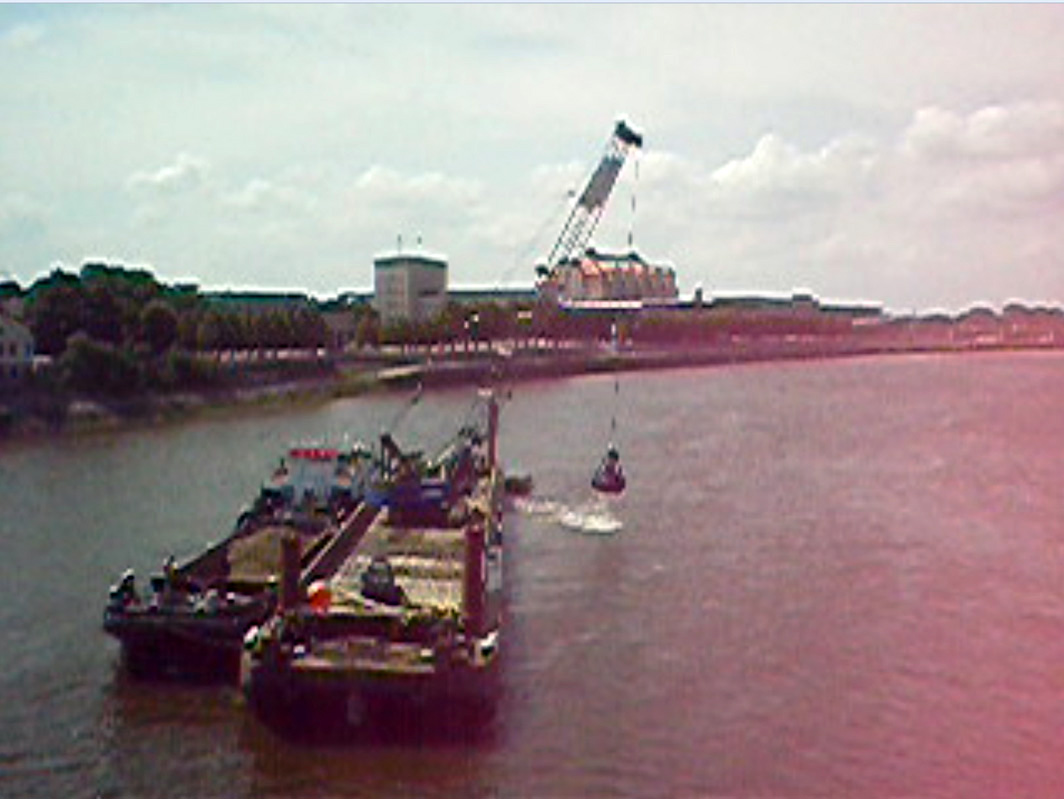 A landscape format, color image that was probably taken from a film due to its poor quality. It shows a very wide river, at the left edge of the picture a bank pulls itself further away into the picture, which disappears at the right edge of the picture. The sky is grayish-cloudy. In the left foreground of the picture are two ships right next to each other, in the middle of the river. On the right one there is a large crane, in the left one there is a large pile of rubble unloaded. The crane has on a rope a dredge shovel hanging just above the water, slightly swinging to the left and from which water is just running.