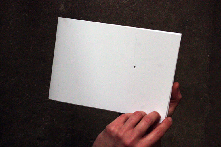 The first of five landscape photos that shows a book from above in the hands of a person. In the background is a dark cement floor. It is white and about 15 by 20 cm, a landscape format. On this first picture is the title page, which is completely white except for a small question mark in the center right.
