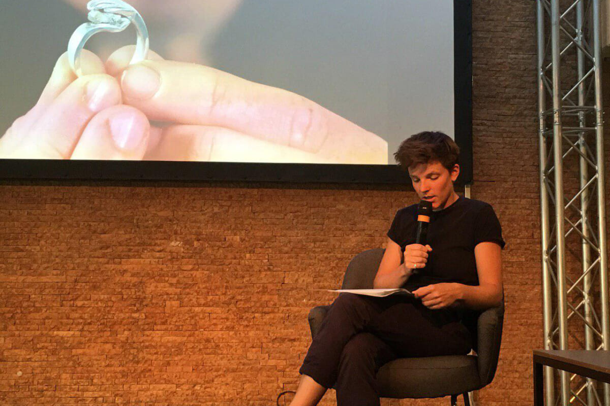 Landscape image of a person sitting on an armchair, dressed in black and holding a microphone. She looks intently at the white piece of paper she has on her lap. She is wearing short brown hair. In the background, on a stone wall, a large canvas is cropped, showing two hands holding a silver ring, the face behind it is blurred.