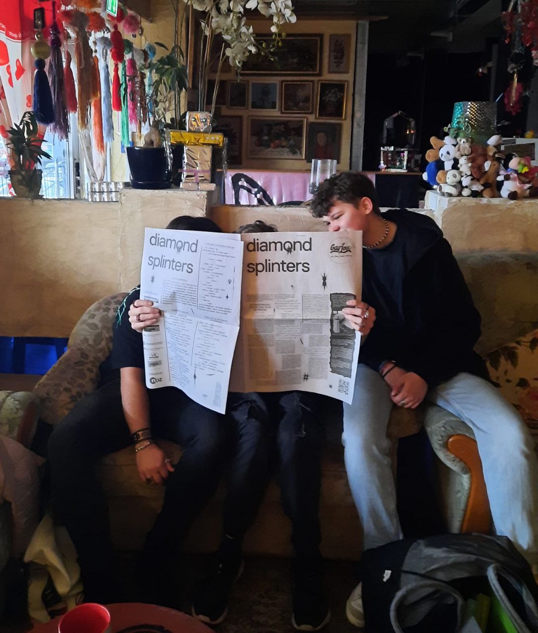 Rectangular color shot of three people in a cozy room, of which, with the exception of the right, the faces are hidden by a large, opened newspaper. It is titled "diamond splinters" and is black and white. The middle person holds it on both sides, only the brown, short hair looks out over the newspaper, from the two left persons only dark jeans and an arm with a leather bracelet are to be seen.