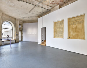 View of a bright showroom with light gray, small checkered tiled floor and two white walls on the right and unrenovated tan ceiling and walls with large round windows on the left. At the very back of the room an entrance door made of glass. On the white wall that runs into the picture from the right hang two large canvases, about 2 meters high, with two archive documents on them that look like scans and show small, black writing on a light brown background. Headphones hang from the ceiling in front of them. Just behind the pictures is a passage in the wall with a staircase leading into it. On the next wall, behind the first and leading toward the entrance, are several, white papers at eye level. From the left edge of the picture a kind of bar runs into the picture, consisting of a board that projects horizontally into the room and is supported by a vertical board, both painted purple. 