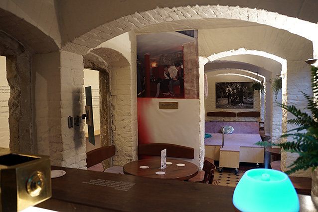 Landscape room view of a cellar vault with white painted brick. The viewer's gaze passes over a wooden bar with a golden tap into the room, which is divided by several round arches. In the first section is a round wooden table with two wooden benches. The wall behind one of the benches is taken up by a photo wallpaper showing a bar scene. A section of the room further back has a built-in pedestal on casters, covered with a lilac carpet. At the very back of the room on the wall hangs a large black and white photo showing three people. In the left half of the picture, a corridor can be glimpsed behind the round arches, with lines of text running along the wall. 
