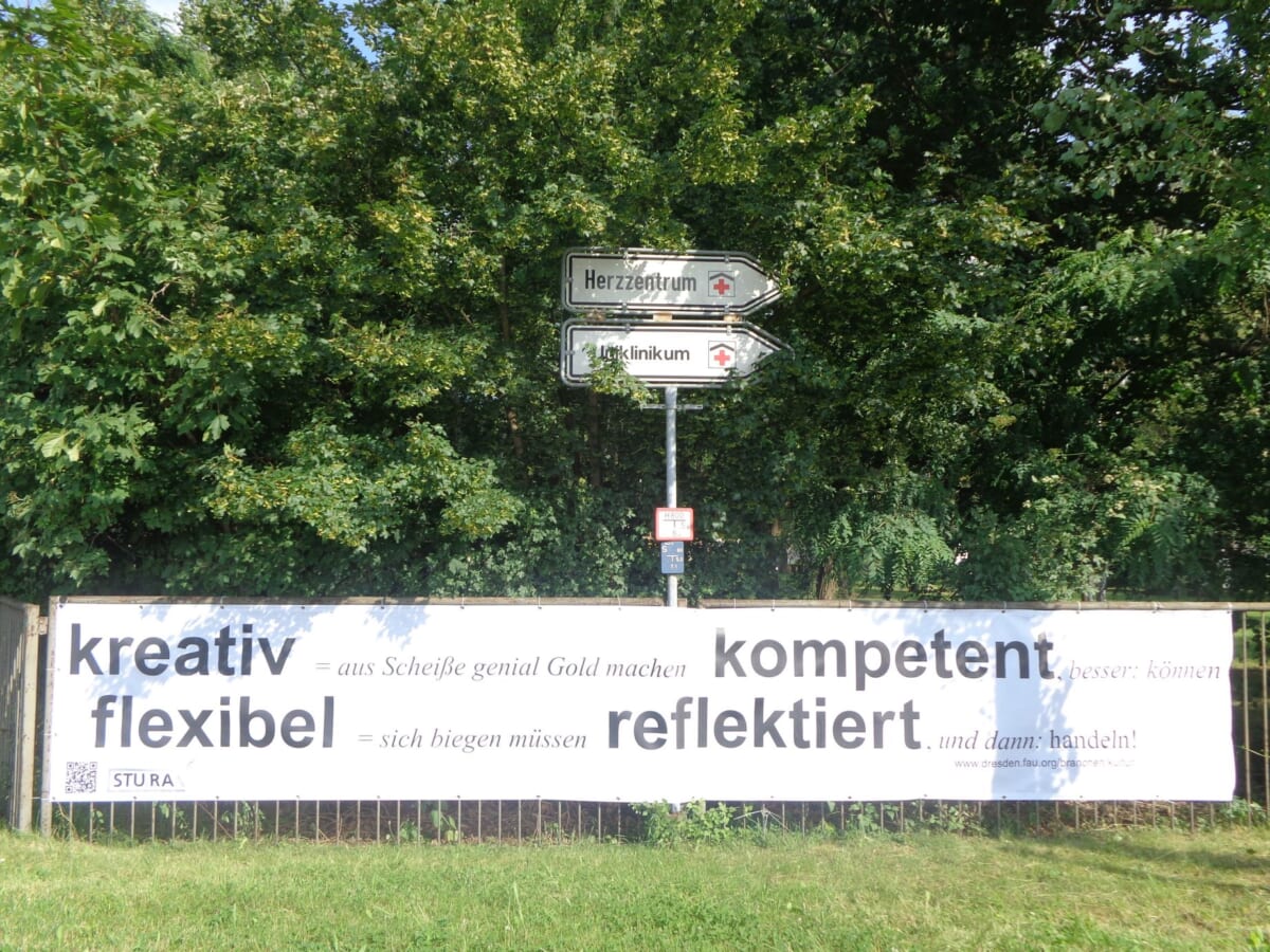 Landscape colored image, the upper two-thirds of which is occupied by densely leafy green trees. In the lower third a low metal fence with many vertical struts, in front of meadow. On the banner hanging on the fence are written in large letters the words "creative, competent, flexible, reflected" and smaller writing that cannot be read. A QR code can be guessed at the bottom left. Above the fence in the middle of the banner is a street sign, the two parts of which both point to the right. It says "Herzzentrum" and " Uniklinikum" on it.