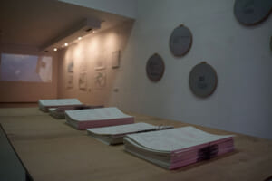 Landscape format color photograph of an exhibition space. In the foreground, a wooden panel on which five stacks of thin paper publications lie, tied with threads whose ends protrude from the stacks. In the background, a room with white walls photographed in the corner, a projection can be seen on the left, 6 large drawings blurred on the right wall and 4 larger, round objects on the front half of the wall. 