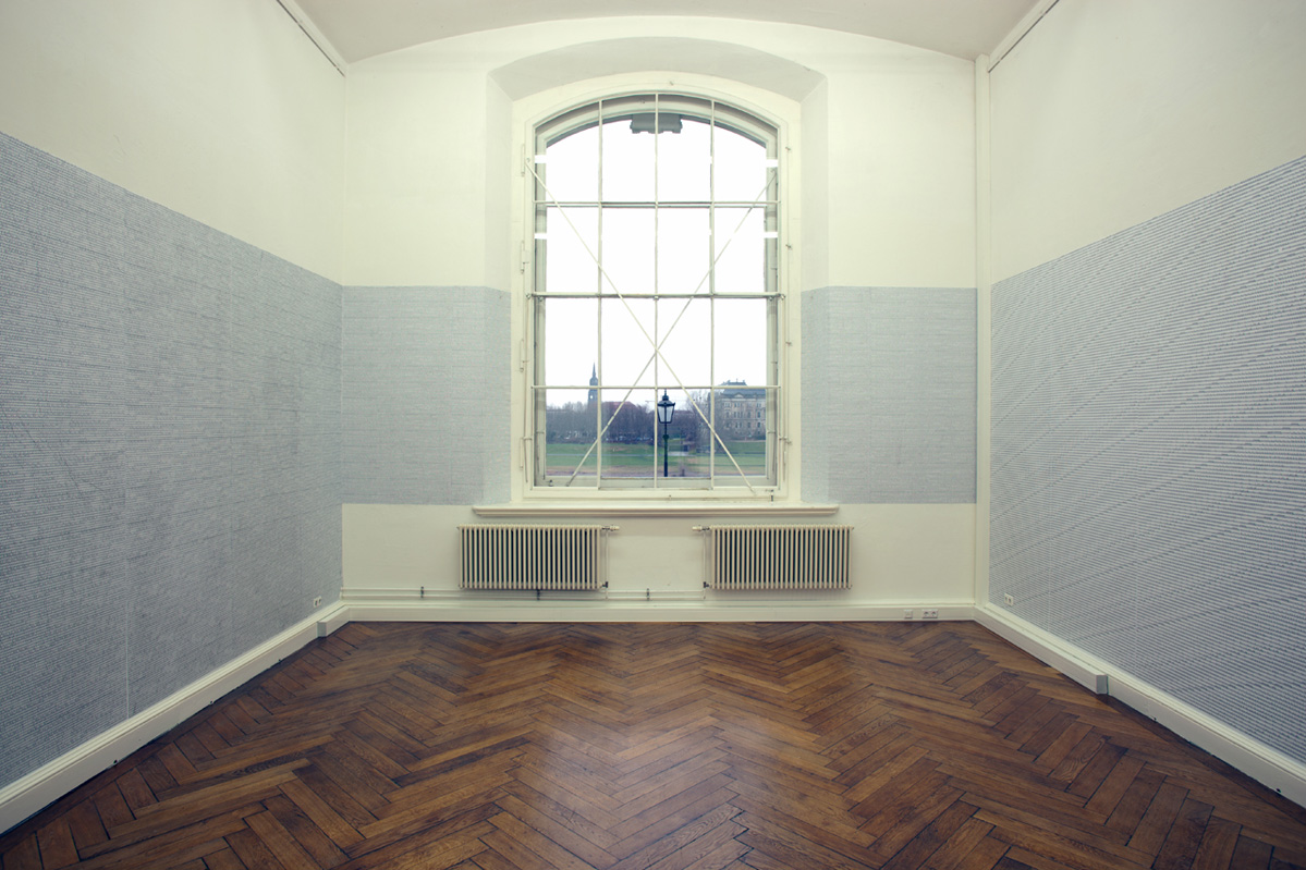 Central general view of the room, photographed towards the wall with a large window. Under the window are two radiators. The right and left walls are covered over and over with small lines of letters up to a height of about 4 meters, sections to the left and right of the window as well.