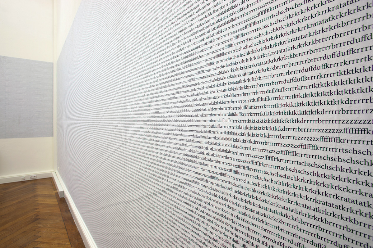 View of one wall of the room, strongly photographed from the right. In the crop on the floor the parquet and on the left wall also covered with letters. The lines of letters running into the picture from the right are very regular and form a pattern over the entire surface. The individual letters are recognizable at the very front. You can read there, for example, "schschkrkrkrkratataschsch".
