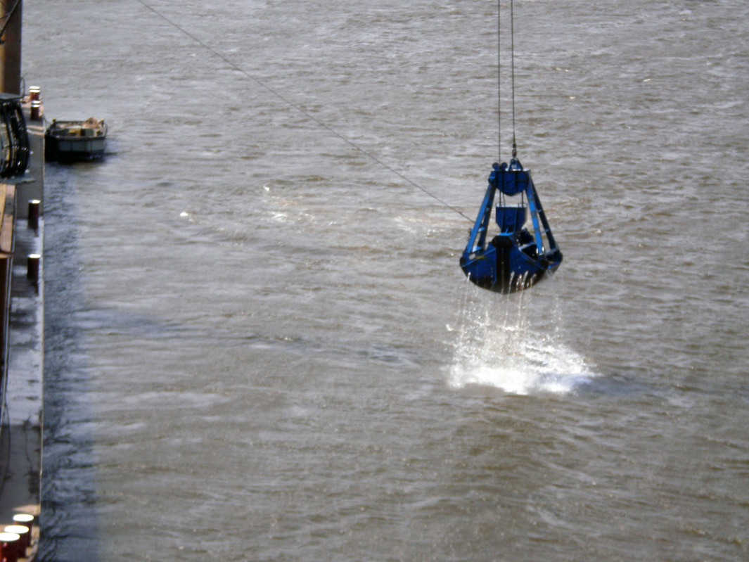 Nahaufnahme der Schaufel über dem Fluss. Am linken Bildrand und ganz leicht angeschnitten das Containerschiff. Das Bild ist sonst eingenommen von der grauen Wasseroberfläche des Flusses mit kleinen wellen. Die Schaufel hängt am Drahtseil wenige Meter über dem Wasser, aus ihr fließt Wasser und bildet weiße Schaumkronen, wo es auf den Fluß trifft.