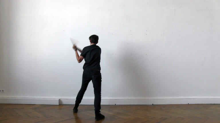Film still: View of a white wall, at the bottom of the picture a dark brown parquet floor. In front of the wall is a person dressed in black with short sleeves and dark, short hair. She can be seen from behind and seems to take a step towards the wall in a quick movement. In her hands, to the left of her body, she holds a stick at forearm length, which she moves quickly and which is blurred. Her left arm is bent to strike.