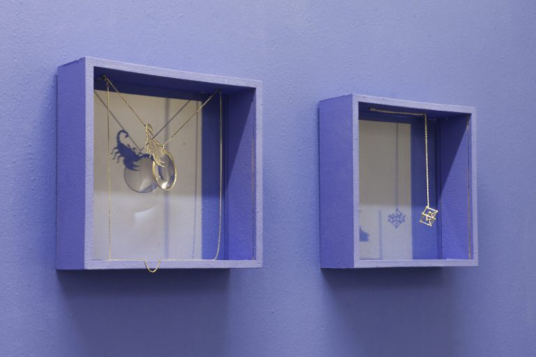 Detail of the two glass display cases, photographed slightly from the left. In the first display case, the chain is clamped between the two upper corners so that the pendant with the scorpion sitting on the glass monocle hangs in the center of the display case. In the left display case, the chain just hangs down to the bottom half of the display case and the pendant is like a chemical structure. The chains are reflected in the glass at the back of the display case.