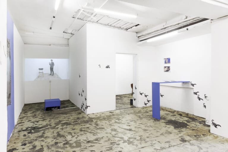 Landscape view of an exhibition room with white walls and ceiling, LED tubes and cables on the ceiling and brown-gray mottled floor. Photographed slightly from the left, the far right part of the room, which is about 50 square meters in size, is separated by more white walls with a passageway. At the right edge of the picture a purple board protrudes from the wall into the room, supported by another one towards the floor. Across several corners, small black scorpions are attached to the white wall of the room at irregular intervals; behind the bar, two photos showing hands hang on the wall above the line of sight. In the back left corner of the room is a projection showing a person in the same room and a chair. The beamer is in a purple box that is on the floor in front of it. In the crop on the right edge of the image is a piece of purple painted wall with a poster.