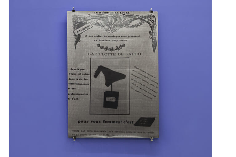 Portrait shot of a purple wall, in the crop below the gray-brown floor. A large black, white and gray poster is attached to the wall with architectural staples and nails. It shows a print of an advertisement that reads "La Culotte de Sapho" in the center, with a small sculpture of a pair of underpants on a pedestal printed in a black frame below. Around the frame is smaller text, at the very top of the ad is a large ornament reminiscent of a vulva.