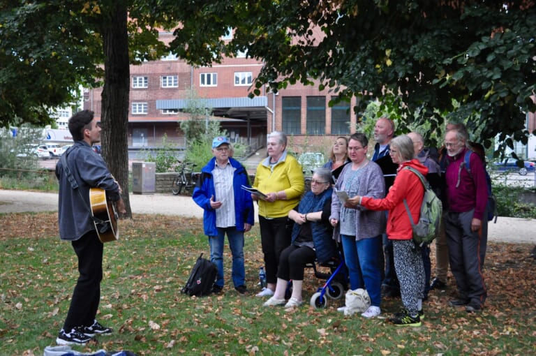 Landscape color photograph of a group standing on a lawn, colorfully dressed and looking into open notebooks, towards a person standing on the left edge of the picture with a guitar. One of the people in the group is sitting on a walker, one has a backpack on, another has a cap on, some have white hair. They seem concentrated. In the background some trees and buildings with red facades.