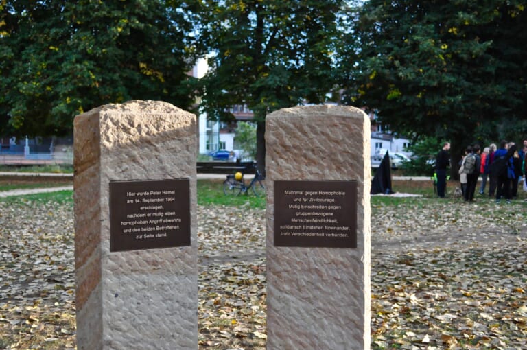 Landscape color photograph showing a lawn in the background, two rectangular sandstone-colored pillars in the foreground, cropped downwards. A brown plaque is attached to the front of each of the columns, the left one reads: "Here Peter Hamel was beaten to death on September 14, 1994, after he courageously defended himself against a homophobic attack and stood by the two victims" and the right one reads: "Memorial against homophobia and for civil courage. Courageously intervening against group-related misanthropy, standing up for each other in solidarity despite differences."