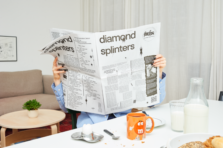Landscape color photo of a person at the breakfast table in a bright room, left in the crop a beige couch, a small wooden table and white curtains. In the foreground on the right a coffee cup, a milk bottle a breakfast egg and rolls. The person is hidden behind the newspaper, which he holds open large and on the front of which is written "diamond splinters ". The newspaper is black and white and various sections of text are set irregularly across the page.