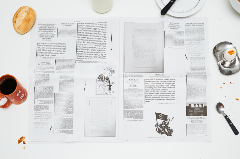 Large format, opened newspaper on a white table, around it in the bleed an orange coffee cup, a roll, a breakfast egg and plate as well as cutlery. The newspaper is printed in black and white with different fonts and irregularly arranged text blocks. In between there are different graphic elements, on the right side there is a black flag with an A in a circle.