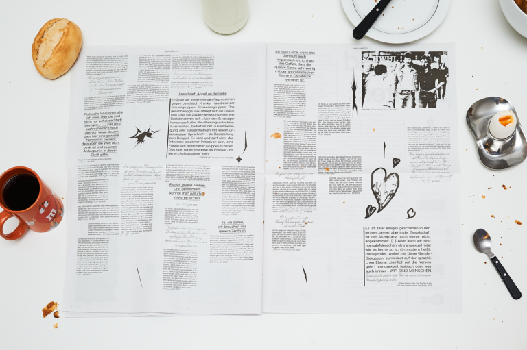 Large format, opened newspaper on a white table, around it in the bleed an orange coffee cup, a roll, a breakfast egg and plate as well as cutlery. The newspaper is printed in black and white with different fonts and irregularly arranged text blocks. In between there are various graphic elements, on the right side there is a black and white photo of two men walking arm in arm at the top and a large hand drawn heart at the bottom.