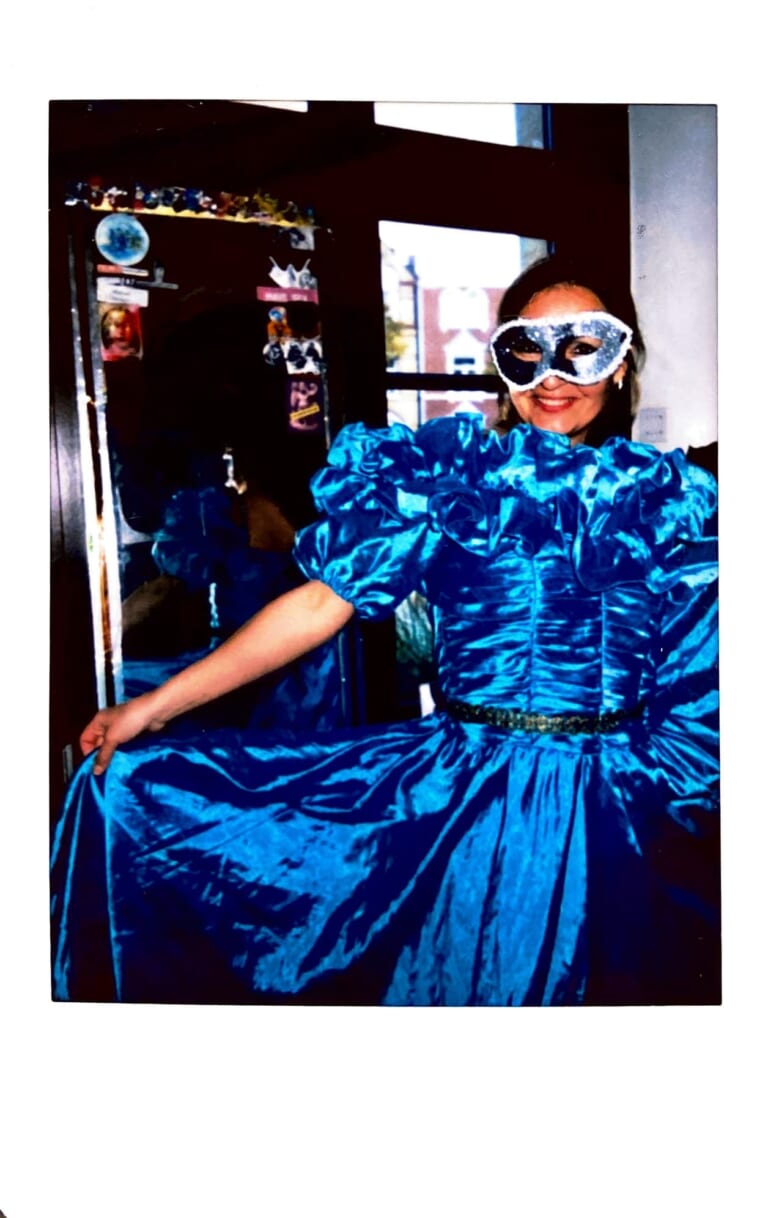 Polaroid photo of a person with long dark hairb wearing a shiny silver mask and a blue satin shiny frilly dress smiling at the camera while lifting one side of the dress with her right arm towards the left edge of the picture. In the background a black door frame and a window.
