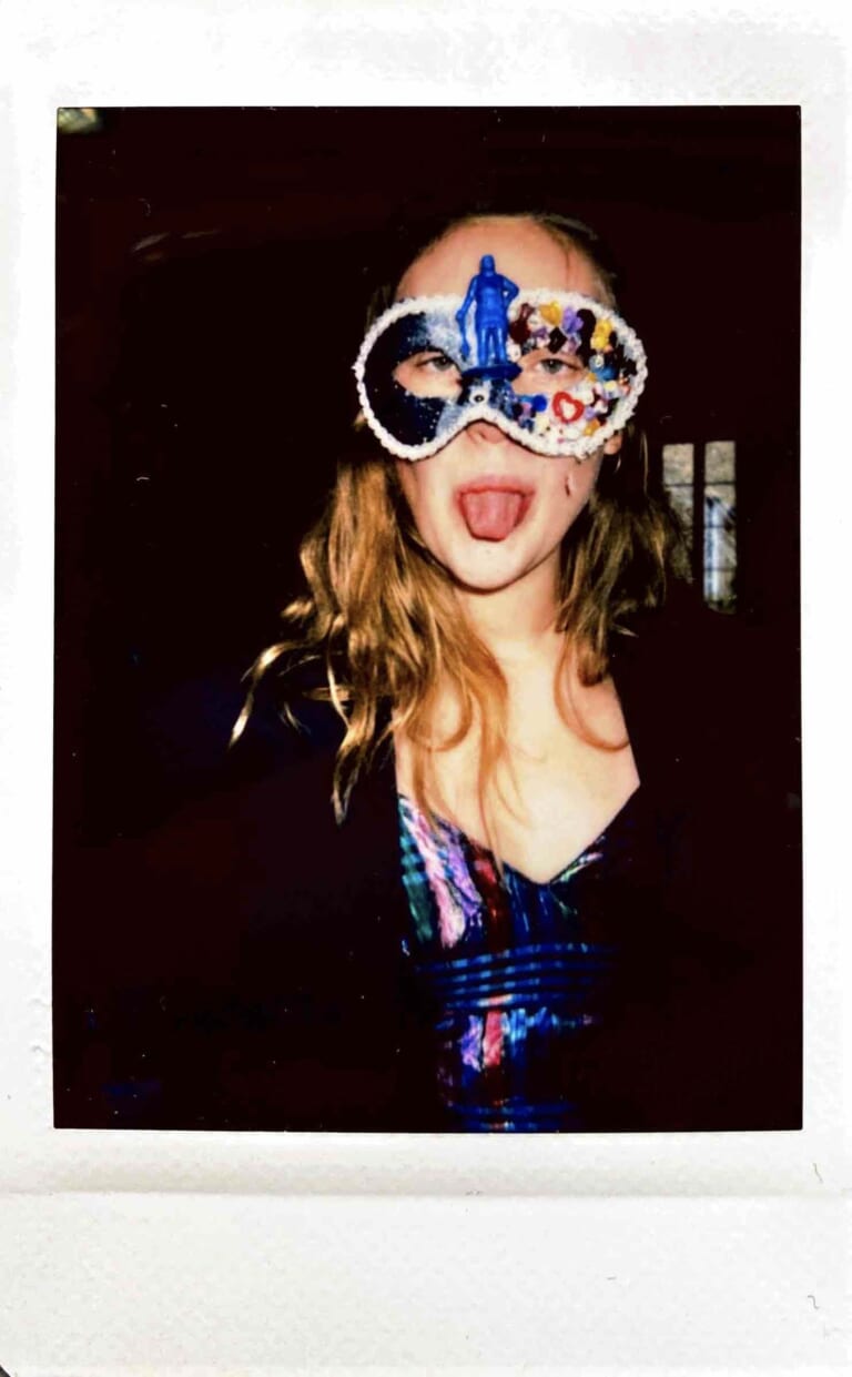 Polaraid of a person from the front, seen from the waist. The background is dark, the person has long light brown hair that falls slightly over the neckline, a black jacket and a colorful shimmering top. She wears a mask with colorful sequins and a blue figure in the nose area. She sticks out her tongue.