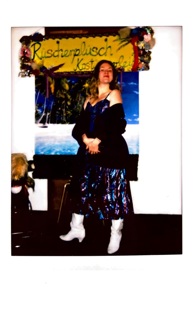 Polaroid depicting a person whole, looking at the camera. At the top of the picture a self-made sign "frilly plush" with colorful decorations and feathers all around. Also, a photo wallpaper with palm tree motif on the wall. In front of it stands the person with white cowboy boots, black shiny strap top and black jacket that fell over his shoulders. The person has his hands folded in front of his belly and looks challengingly into the camera, wears light brown long hair and a colorful shimmering dark skirt.
