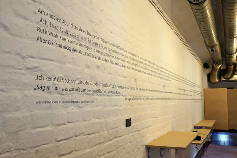 View of a long corridor with white painted brick, photographed from the left wall. Silver pipes run across the ceiling, and in the background the wall opens into a wooden hinged door. At the bottom of the wall are wide wooden boards of various heights of seats. Along the entire length of the wall runs a text with several lines, of which only the beginning is in focus.