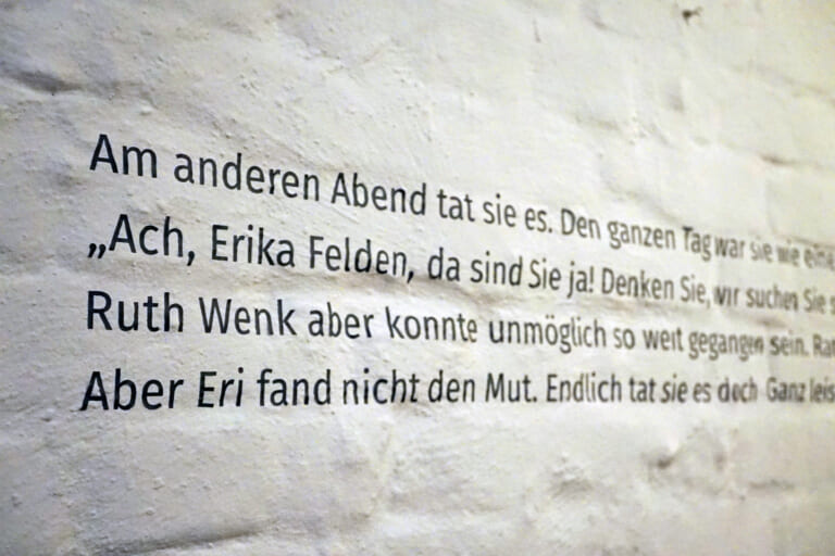 Close-up of the dark gray text on the white painted wall, photographed slightly from the left. To recognize are the words in German"The other evening she did it. ...Ah, Erika Felden, there you are!"...Ruth Wenk, however, could not possibly have gone that far...But Eri could not find the courage. Finally she did..."