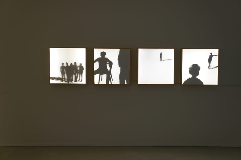 Detail photograph of the wall with the light boxes. The focus is on the shadows of the light boxes, so the exhibition space appears very dark. At the bottom of the image is the baseboard of the room. The lightbox on the far left shows a five people standing in a group casting a shadow to the left. Their silhouettes are partly sharp, partly very blurred. The lightbox next to it shows a seated person with legs spread apart, as well as a standing person who can be seen from behind, looking in the direction of the seated one. On the third lightbox, a small standing person can be seen at the top with their arms crossed and appearing to be smoking. She casts a shadow to the right. The last lightbox in the series shows the back of the head of a person with short hair looking toward a person who is leaning on the right edge with her arm, legs crossed, looking back at the other person. She also casts a shadow to the right.