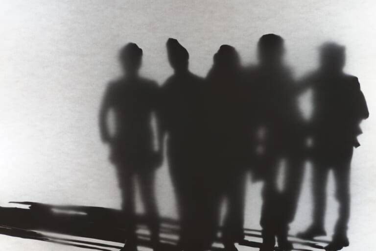Close-up of the group of people from the left light box. The right two-thirds of the image are occupied by the five people, whose silhouettes are partly sharp, partly blurred. The white image space has the texture of paper, the black silhouettes seem to emerge from behind. The persons are facing each other, they can all be seen from the front. Their long shadows run to the left edge of the picture.