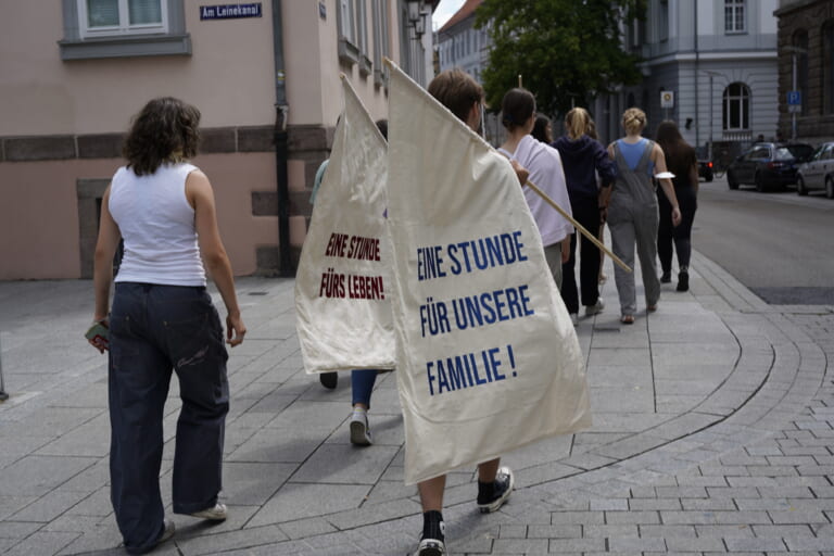 Landscape color shot of a group of people from behind, walking on a sidewalk past a house on the right. They wear long pants and partly T-shirts, on the right image frame in the crop another house and cars. The group is carrying two flags made of beige fabric on wooden flagpoles that are almost as tall as the people themselves. One of them reads in blue "One hour for our family!", the other one in red "One hour for life!
