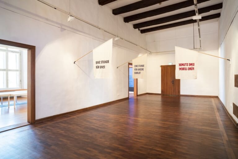 Landscape color shot of a large room, photographed slightly from the back right corner into the room. Dark shiny parquet, white walls and ceiling structured by dark wooden beams. On the right wall, two open doors lead off into other rooms. Between these two doors hang two beveled flags with writing and beige fabric, and on the right wall hangs another. The flags read in black, red and blue letters: "One hour for us! One hour for our family! One hour for life!"