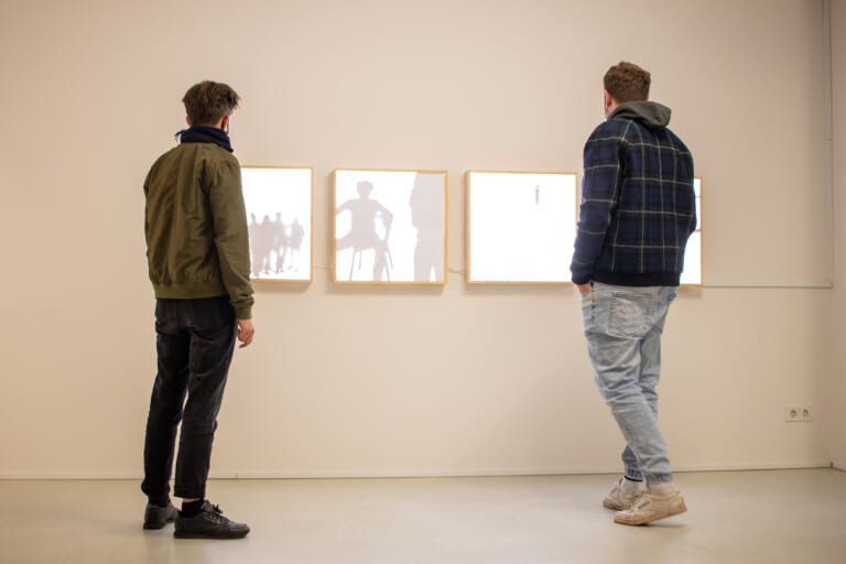 The landscape color photograph shows the white wall on which four light boxes hang. To the left and right at the edge of the image are casually dressed people looking in the direction of the light boxes. The light boxes show black, shadowy silhouettes and are mounted slightly below viewing height. They have wooden frames. The lightbox on the far right is obscured by the person looking at it in the room.
