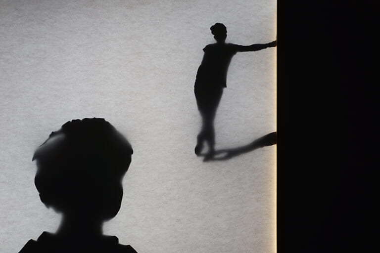 Another detail of the right light box. It shows the back of the head of the person with short hair looking towards a person who is leaning on the right edge with his arm, legs crossed, looking back at the other person. She also casts a shadow to the right. Here, too, the structure of paper is recognizable, the silhouettes are partly sharp, partly blurred.
