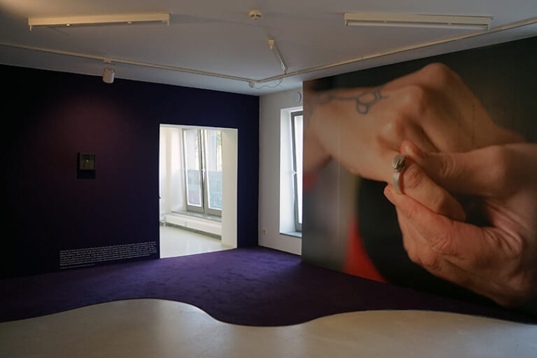 Landscape color photograph of the exhibition space, photographed in the corner with door and the long window. On the right side the wall with the photo wallpaper of the ring protrudes into the picture, the left wall is painted dark purple, in front of it snakes along the carpet in the same color. On this wall, to the right of the door at foot level, is a block of text in white lettering, above which is a small display case at eye level, also painted purple. A very small silver ring is visible behind the glass pane.