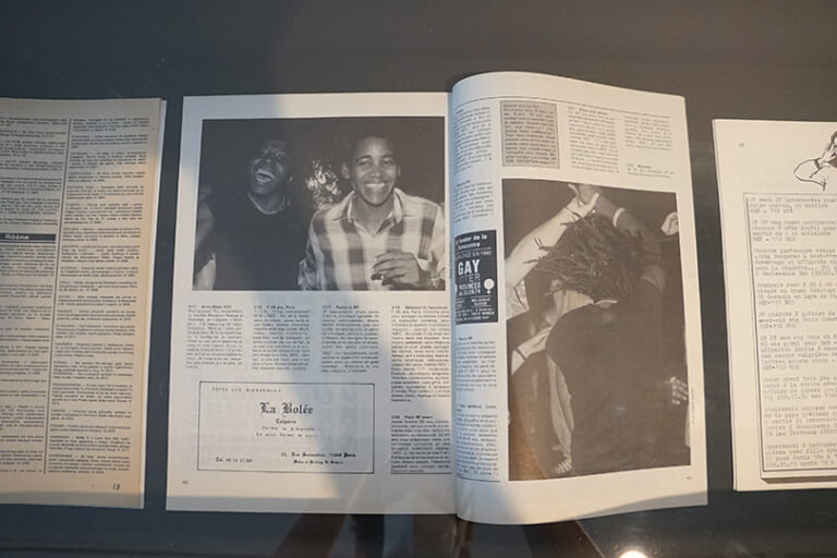 Another detail shot of a magazine from the showcase, also black and white. The left page shows a photo of two black women, laughing broadly, looking at the camera. They are in a party mood, wearing short hair, the one on the right a plaid shirt. Below them are classified ads in French, and an ad for a bar called "La Bolée" is also printed. The right side of the magazine shows more ads and a large photo of another black person with dreadlocks spinning in circles while dancing.
