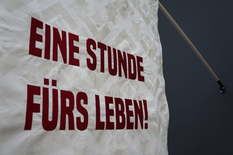 Landscape color close-up of the beign textile filling the left three quarters of the image. On the right side the flagpole holding the fabric protrudes into the picture, the wall it is attached to is black. On the fabric, which has a light pattern with large triangles, is written in large red block letters "An hour for life!".