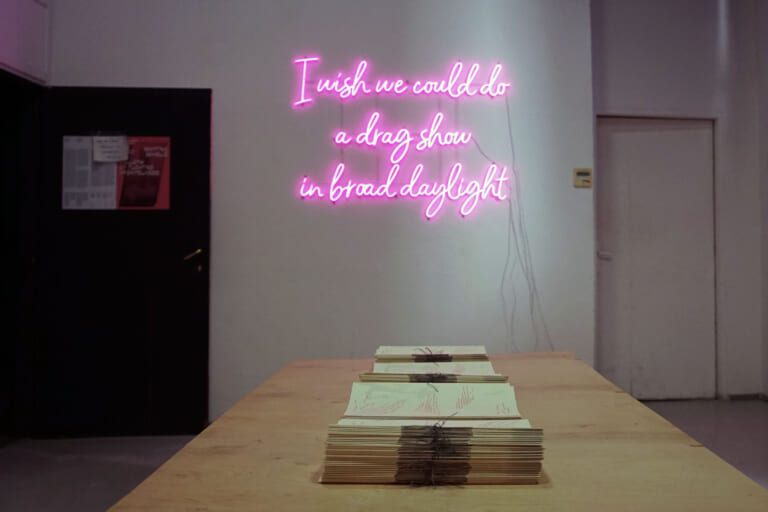 Photograph of the exhibition space, on the left a black door, on the right a white door that is closed. On the wall, a frontal view of a pink neon sign that lights up: "I wish we could do a drag show in broad daylight". In the foreground, a wooden tabletop with stacks of paper publications.