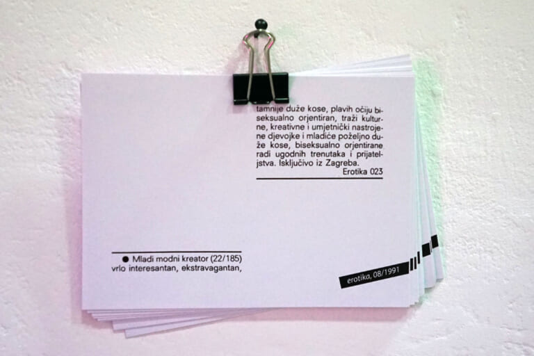 Close-up of a stack of white postcards held together with a staple and hanging on a nail. Two blocks of text are printed on the front postcard, which can be recognized as a personal ad in Croatian. A small black bar at the bottom right reads "erotika, 08/1991".