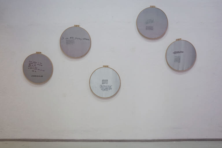 Landscape view of a white wall to which five large, round embroidery frames are attached at different heights. Silver fabric is stretched in them, small blocks of printed text resemble the contact ads on the postcards, handwritten lines are embroidered next to them in different colors.