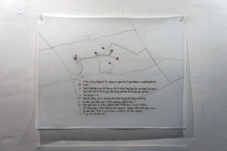 Another close-up of a cartographic drawing. Pencil lines can be seen on the back of the paper, which repeatedly run towards some purple dots. A handwritten text about queers at the university and on campus is written at the bottom center of the tracing paper, which is attached with silver paper clips.