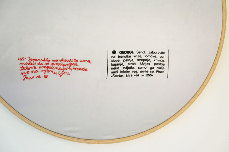 Close-up of an embroidery hoop with silver fabric. On the right is a printed personal ad in Croatian, beginning with the words "George Sand", on the left a small embroidered red reply ending with a small heart.
