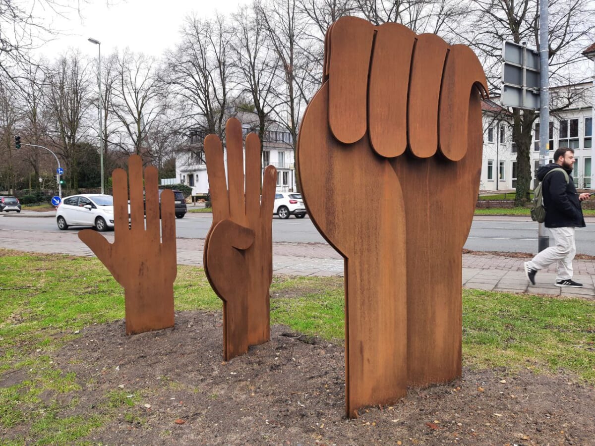 Landscape format color photograph in a public space. A street in the background, a lawn in the foreground, on which three hands made of rusty metal stand in a row. The smallest one, at the back, is flat, like a "stop" sign. The middle one shows a folded thumb, the largest in the front a fist.